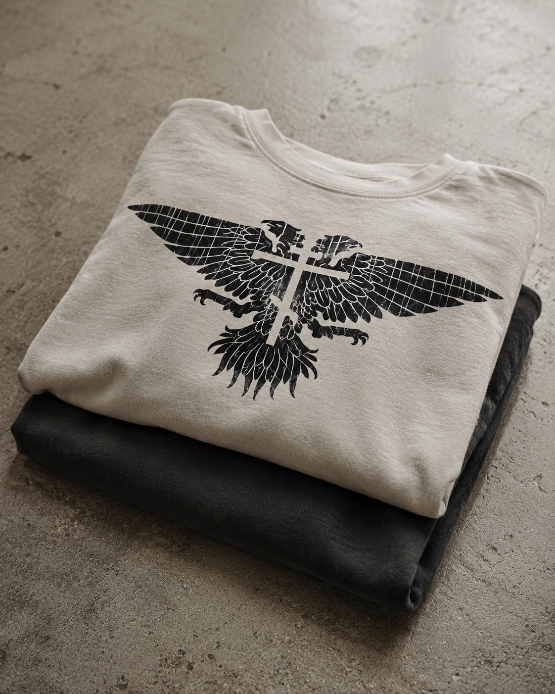 A folded off-white t-shirt with a black graphic of a winged cross and eagle, resting on top of a folded black t-shirt on a textured concrete floor.