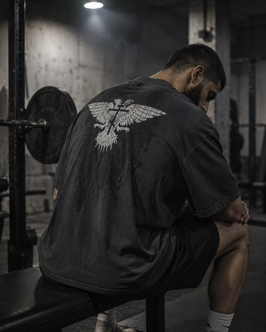 A man sitting on a gym bench viewed from behind, wearing a dark gray t-shirt with a large white double-headed eagle graphic on the back. The gym environment is dimly lit with concrete walls and weight equipment visible in the background.